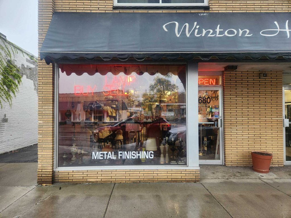 A neighborhood shop with a dark blue “Winston” awning, glowing “Buy Sell” and “Open” signs, and metal finishing text on the window.
