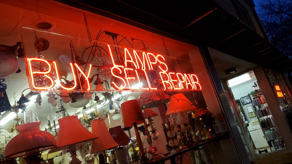 A vintage lamp shop window glows with red neon reading “Buy, Sell, Repair,” surrounded by lamps and fixtures, creating a warm, nostalgic street scene.