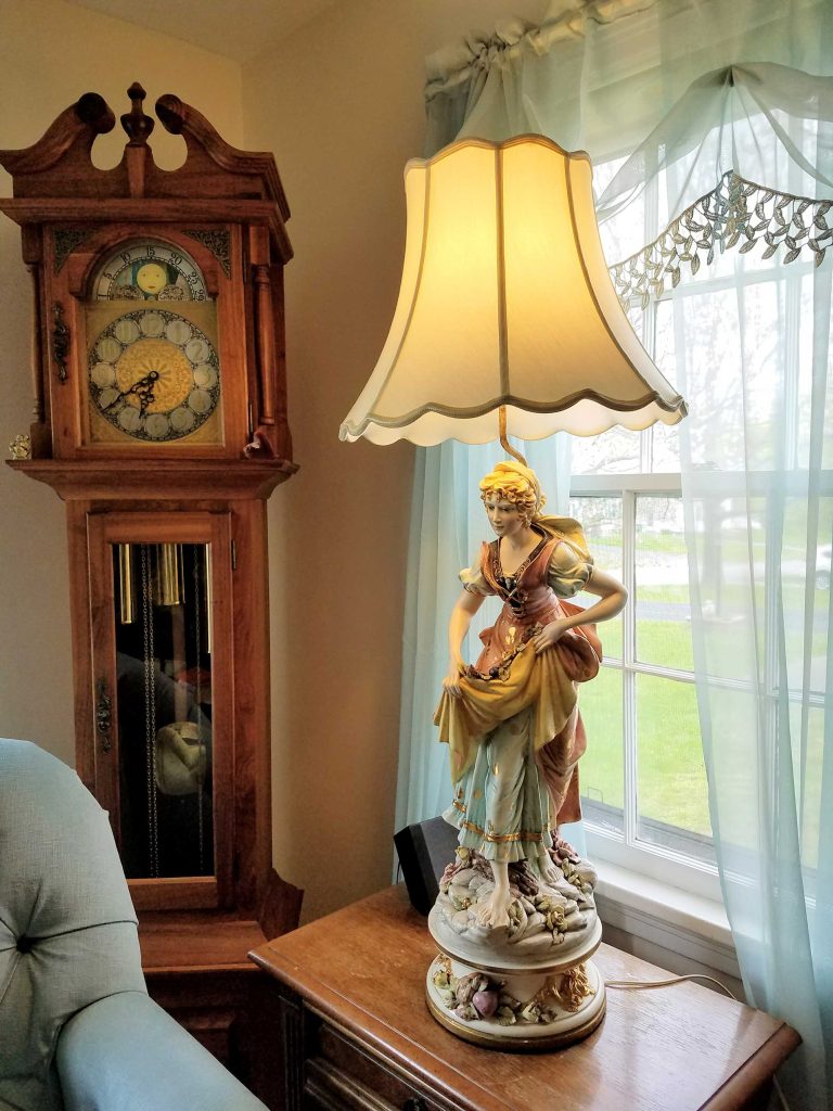 A warm vintage sitting room with a carved wooden grandfather clock, glowing figurine-base table lamp, blue armchair, and bright curtained window.